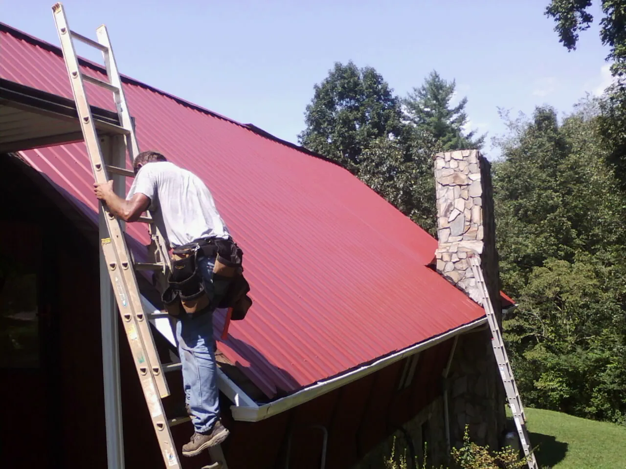 Roofer on a ladder beside a red metal roof and stone chimney — Soffit & Fascia Repair by Roofing Brothers in Shaniko